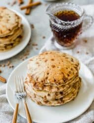 A stack of cinnamon chip pancakes on a plate with maple syrup in a clear glass in the background.