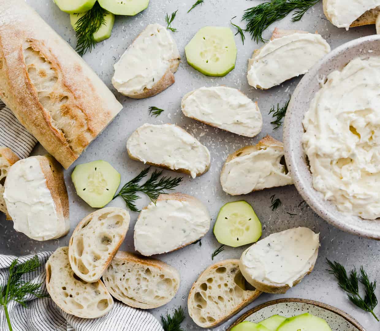 Ranch cream cheese mixture smeared atop sliced baguettes, with cucumbers in the background.