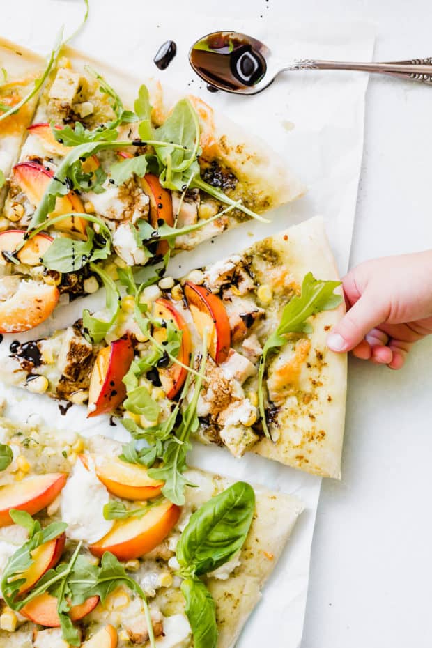 A Childs hand grabbing a slice of pizza that is topped with chicken, nectarines, arugula, and a balsamic drizzle. 