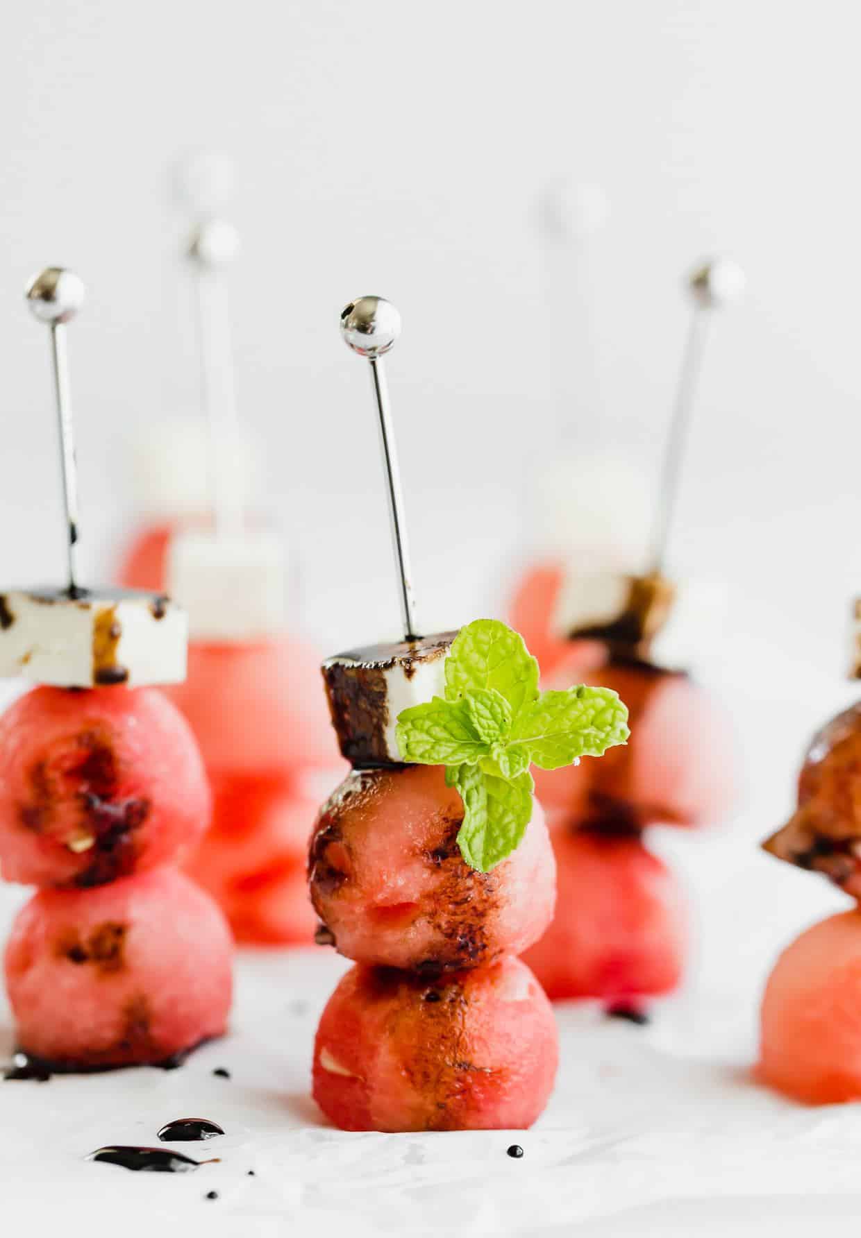 Two watermelon balls and a block of feta on small skewers, drizzled with balsamic reduction and a mint leaf for garnish.