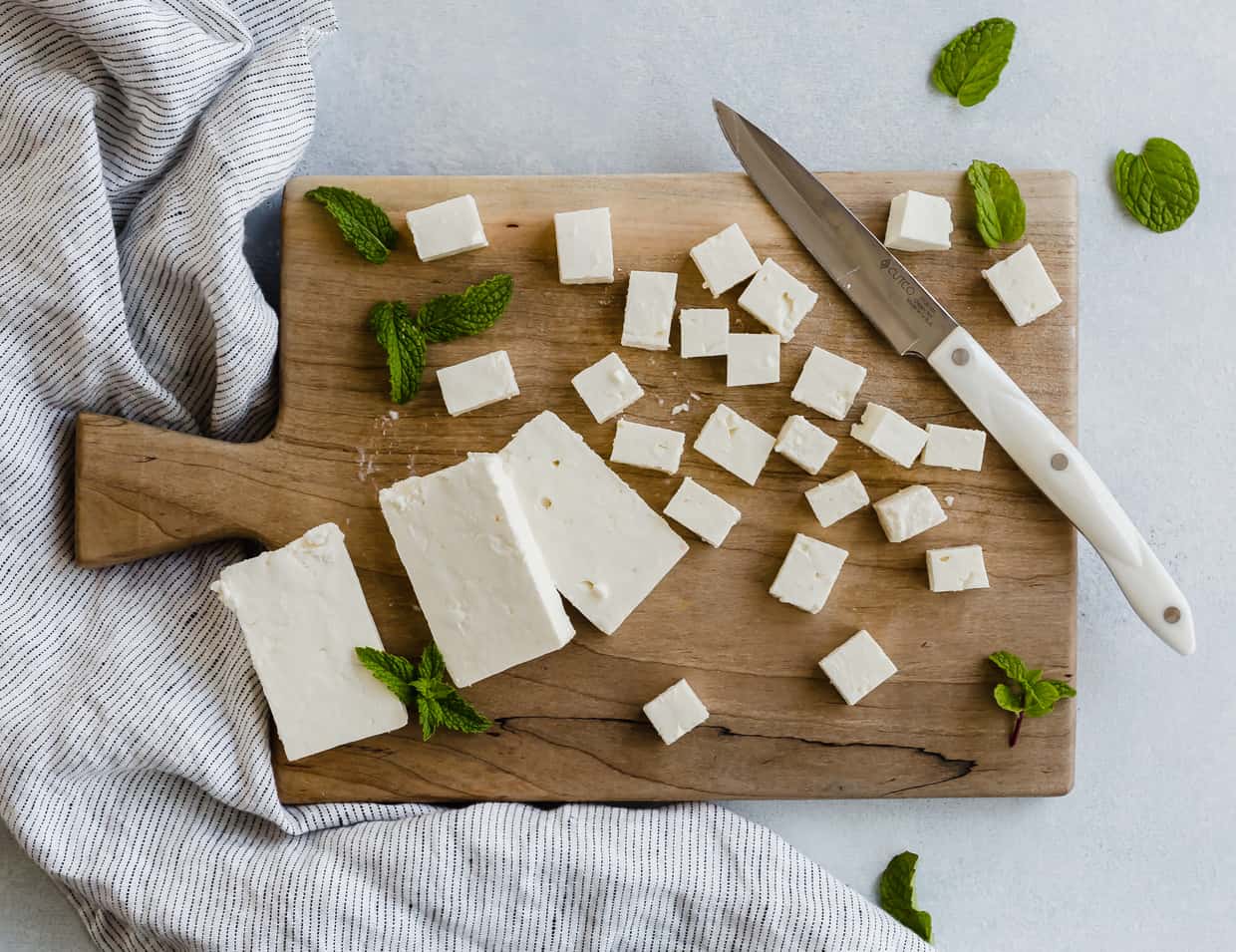Overhead view of a wooden cutting board with a block of feta cheese cut into small squares.