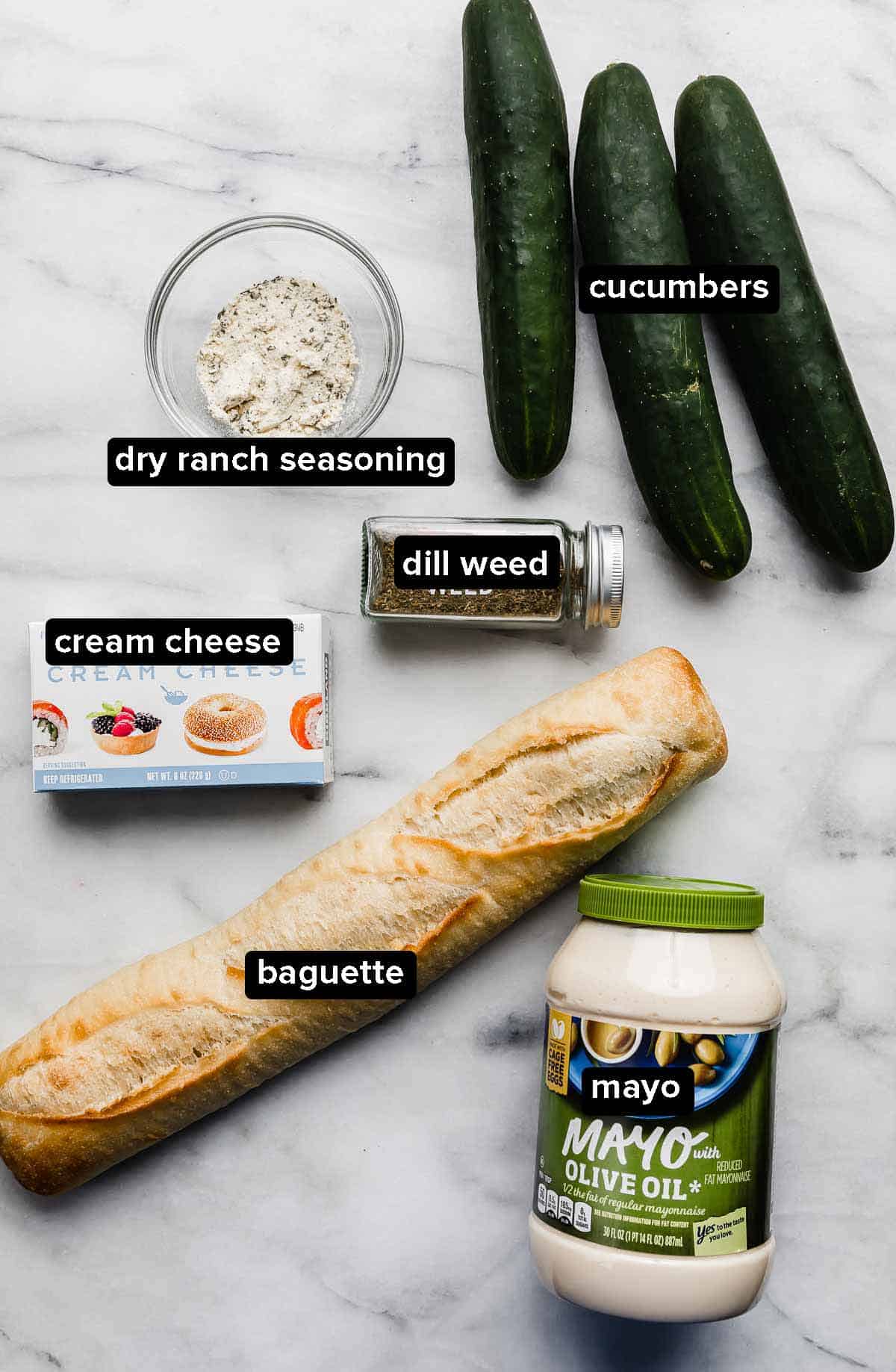 Cucumber Sandwich Appetizer ingredients on a white background: dill, cream cheese, cucumbers, baguette, and mayo.