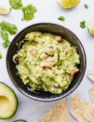 Bowl of guacamole surrounded by tortilla chips, cilantro, and limes.