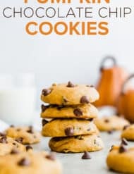 A stack of 4 pumpkin chocolate chip cookies and 2 pumpkins in the background.