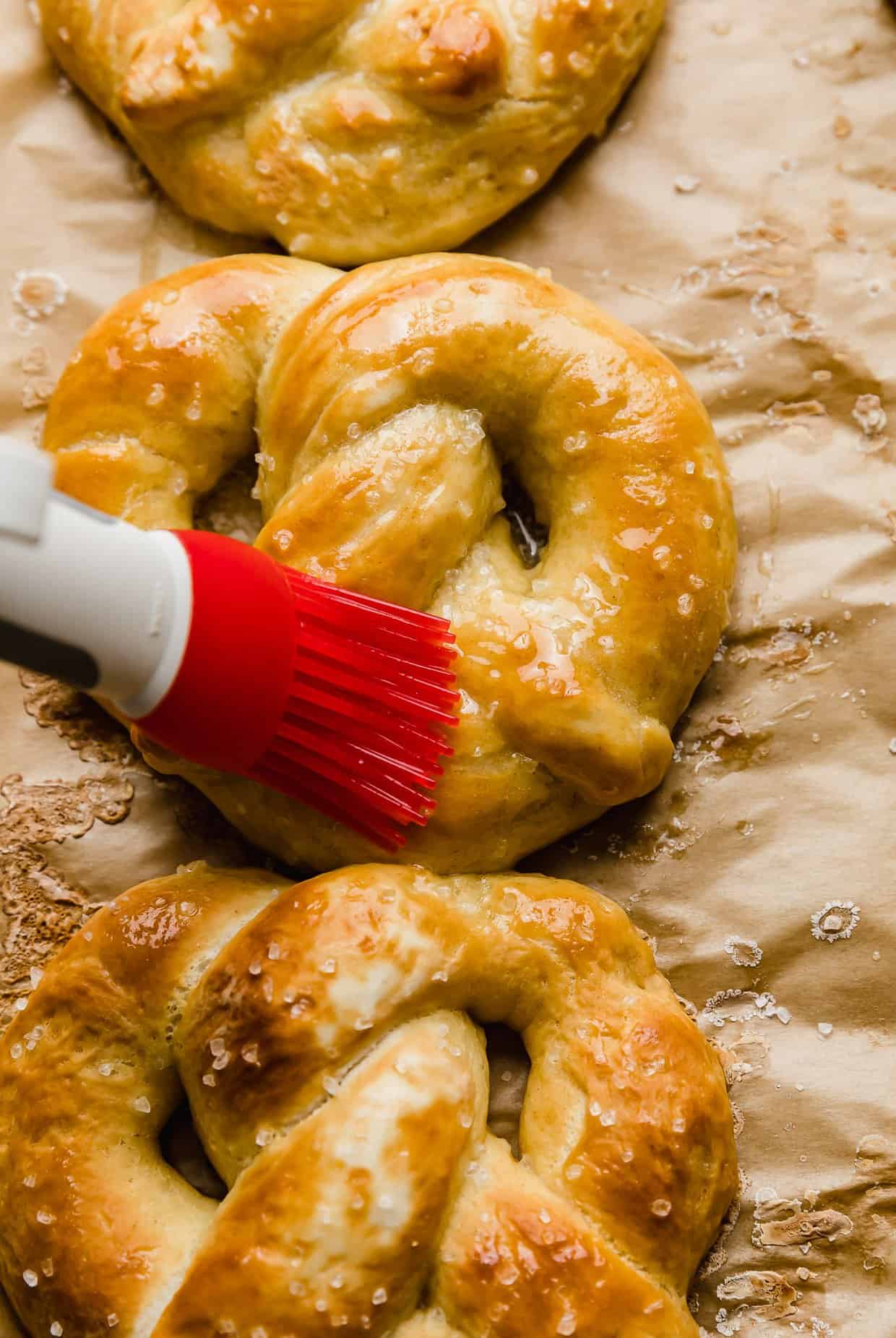 A red pastry brush spreading melted butter over a golden baked pretzel.