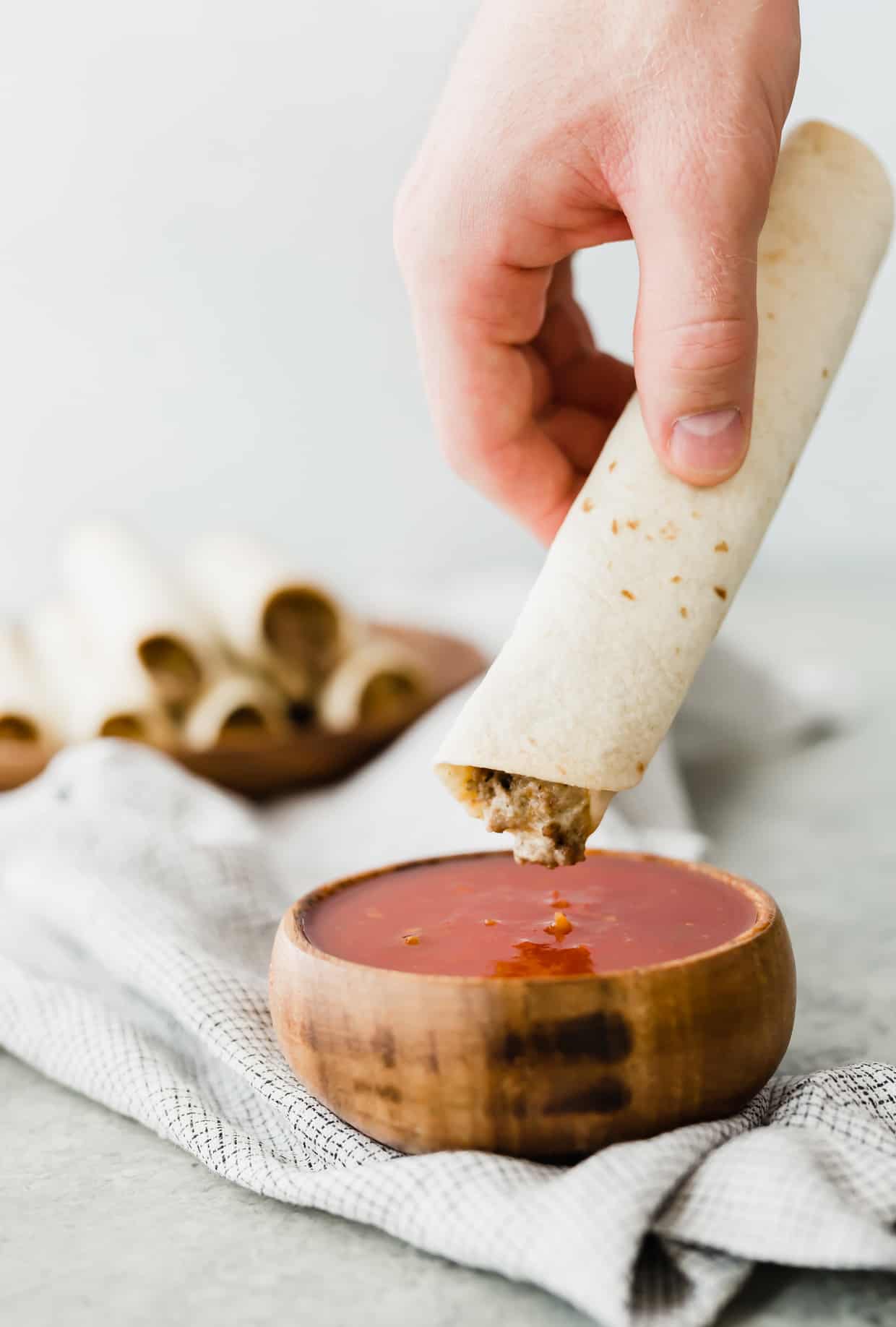 A hand dipping a taquito into a bowl of red salsa. 