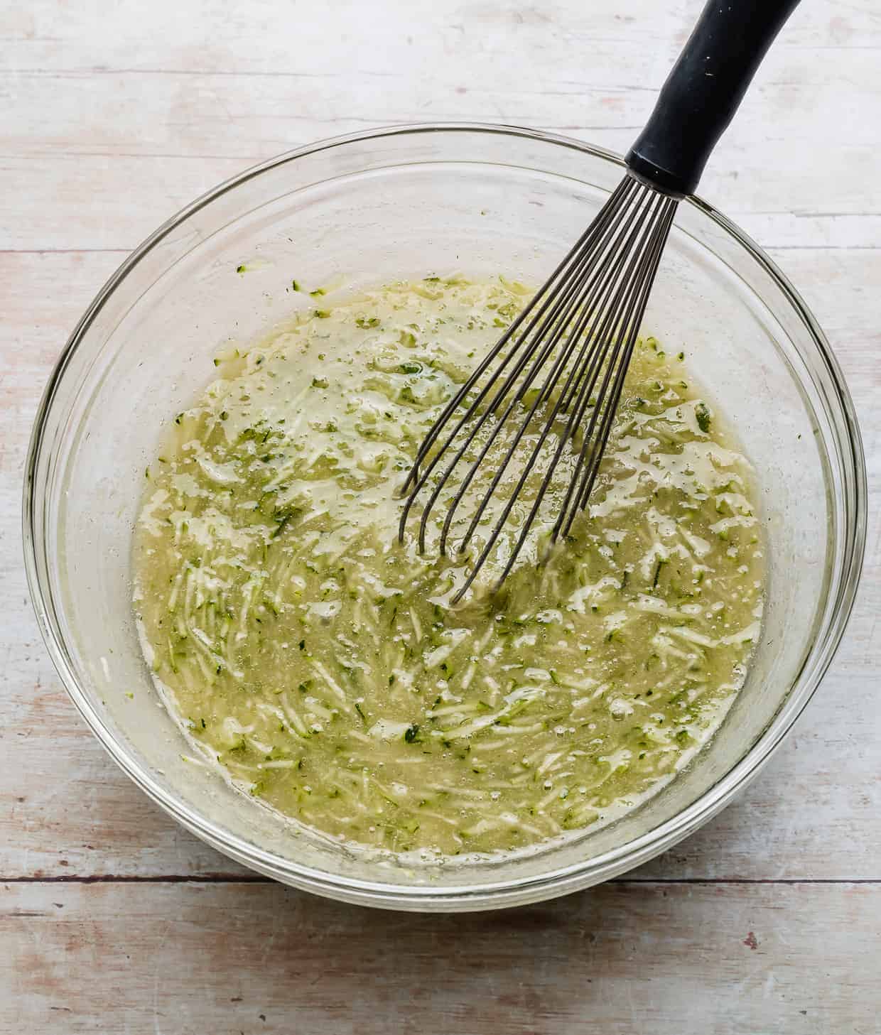 Shredded zucchini in a glass bowl with a whisk.