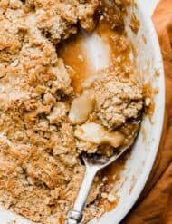 A serving spoon scooping out homemade apple crisp from a white baking dish.
