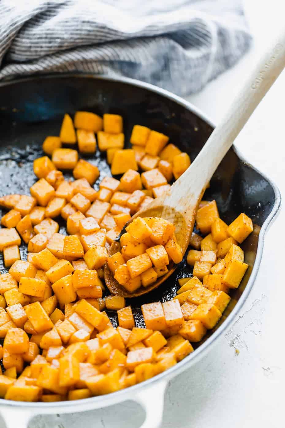 A wooden spoon scooping up diced butternut squash from a skillet.