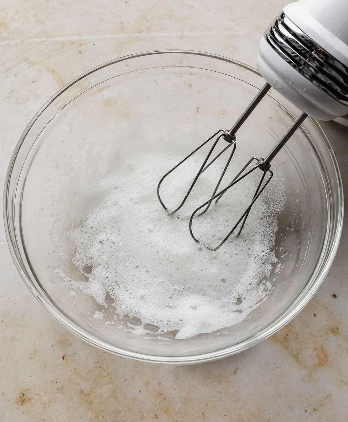A hand mixer whipping egg whites until frothy in a glass bowl.
