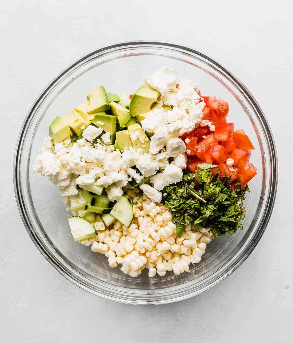 A glass bowl full of corn kernals, diced tomatoes, feta cheese, avocado and mint.
