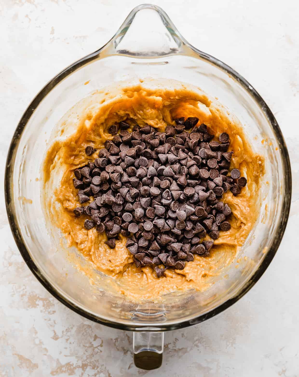 Chocolate chips overtop pumpkin chocolate chip cookie dough batter in a glass bowl.