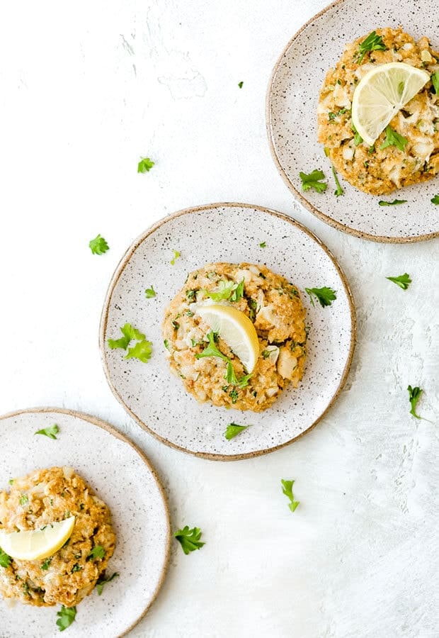 Three tilapia cakes on 3 different plates with a lemon wedge atop each.