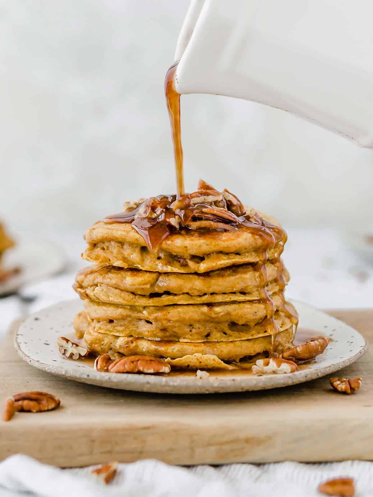 Pumpkin pancakes with pecans on top and syrup being poured over the pancake stack.