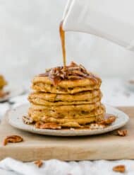 Fluffy pumpkin pancakes topped with pecans with syrup being poured overtop of the stack.