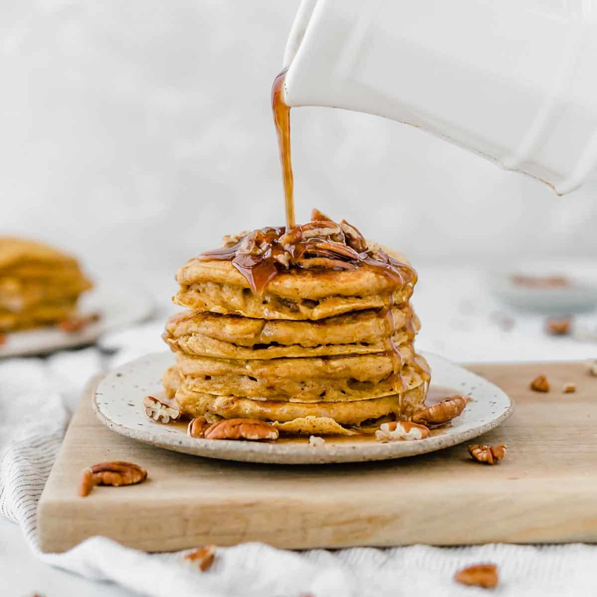 Fluffy pumpkin pancakes topped with pecans with syrup being poured overtop of the stack.