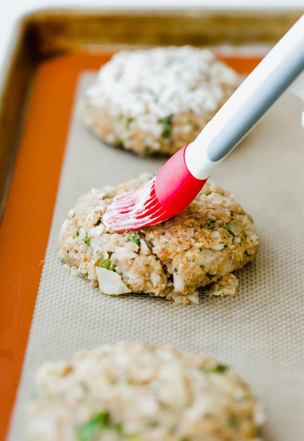 Tilapia cakes on a baking sheet with a pastry brush spreading mayo overtop.