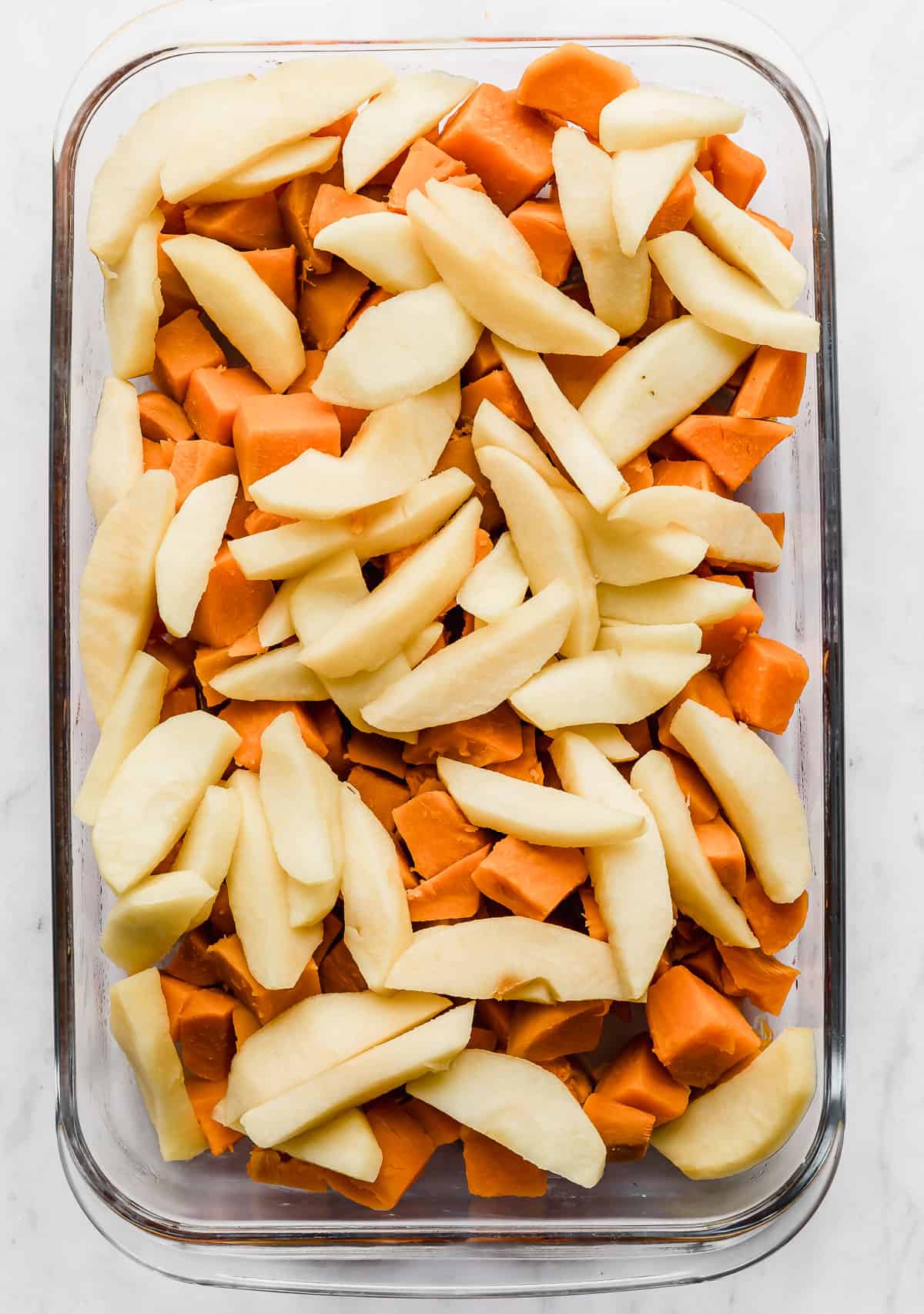 Sliced apples overtop cubed sweet potatoes, in a glass casserole dish.