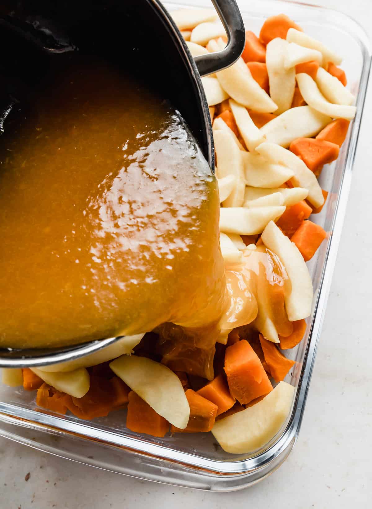 An orange juice sauce being poured over cubed sweet potatoes and sliced apples in a casserole dish.