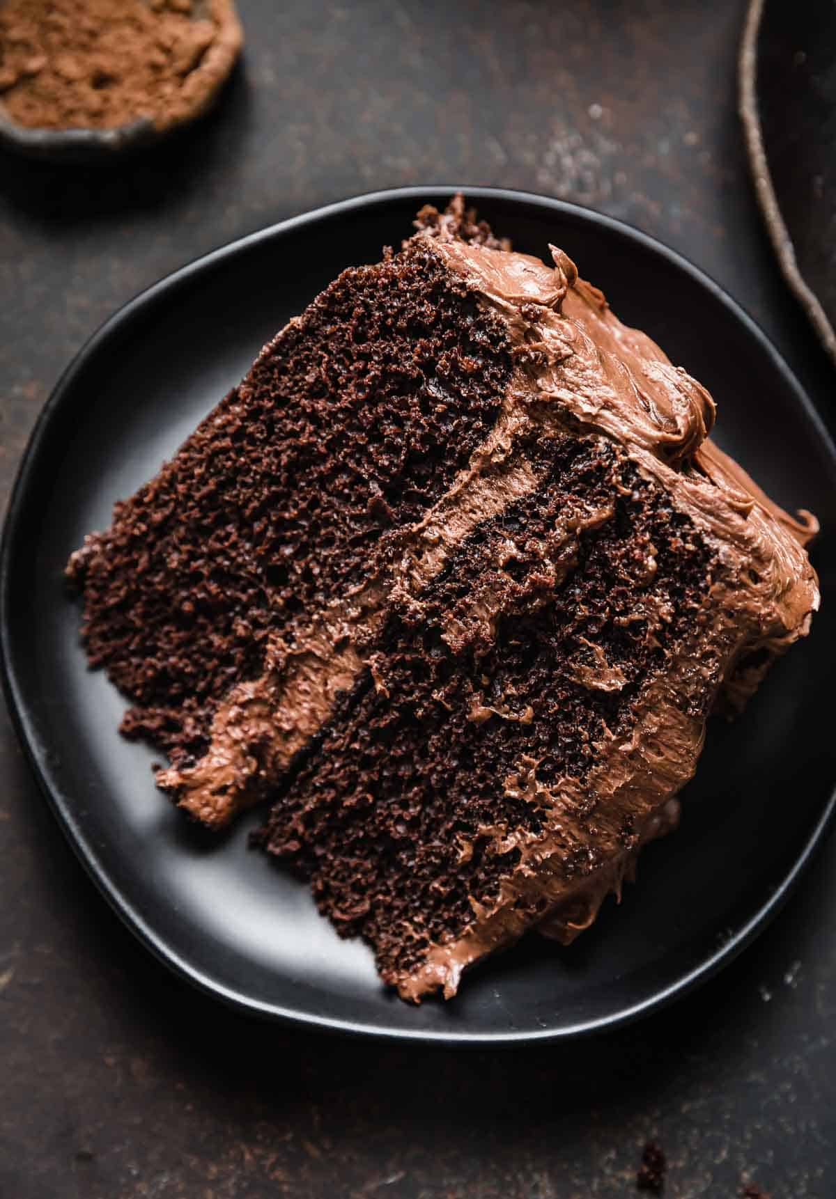 Two layer chocolate cake with a decadent chocolate frosting, on a black round plate.