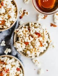Overhead photo of a bowl of maple bacon popcorn with popcorn kernels scattered around the bowl and a bowl of maple syrup in the top corner.