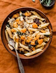 A brown bowl filled with Butternut Squash Mushroom Pasta that's topped with parmesan cheese, all on a burnt orange linen tablecloth.