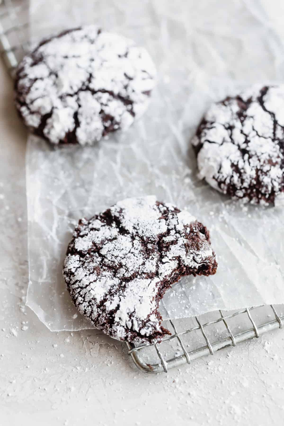 A Chocolate Crinkle Cookie with a bite taken from it, on a metal cooling rack on a white background.