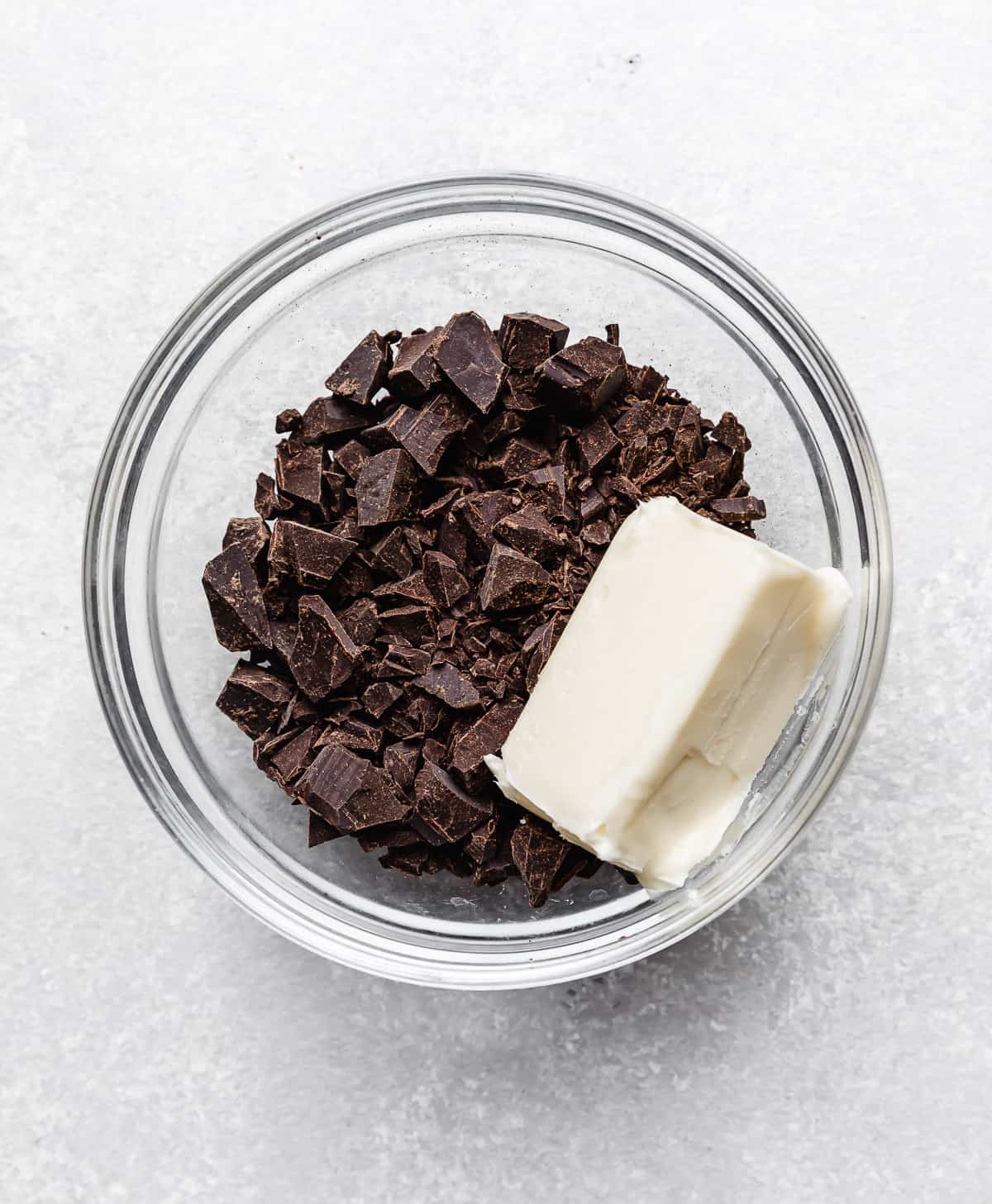 A stick of butter and chopped chocolate in a glass bowl on a light gray background.