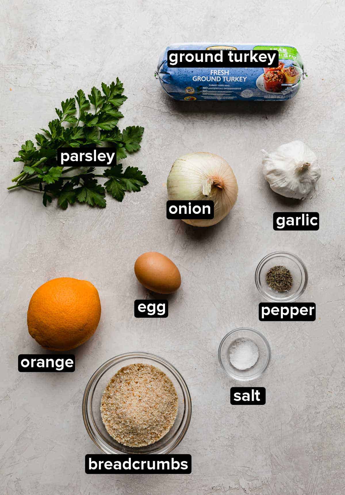 Christmas turkey meatballs ingredients on a light gray background, which include: parsley, orange, breadcrumbs, egg, onion, and turkey.