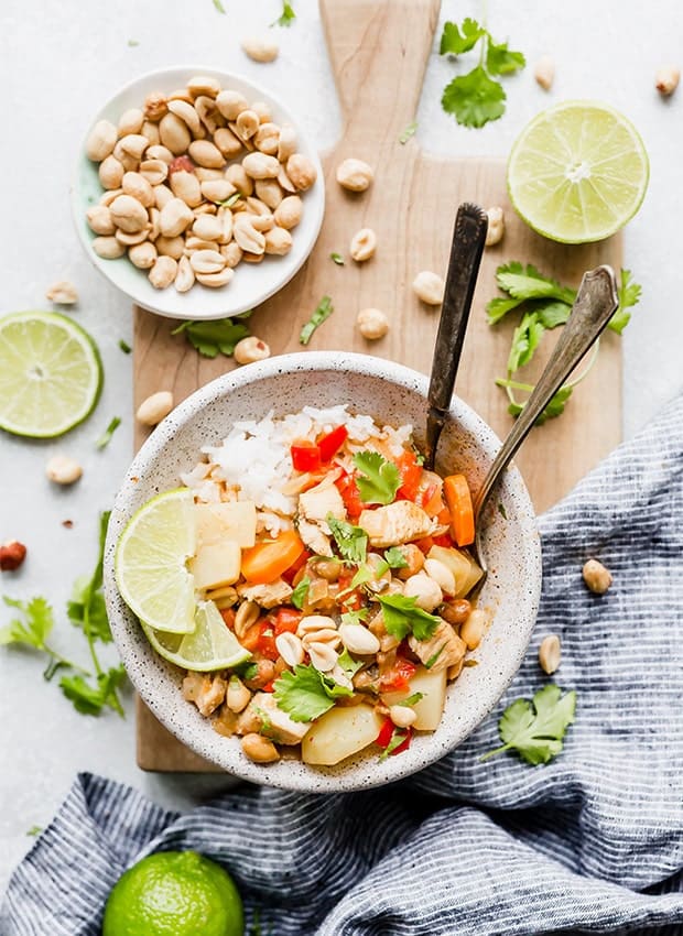 A bowl full of white rice and Massaman curry set on a wooden cutting board with cilantro, limes, and peanuts scattered around the board.