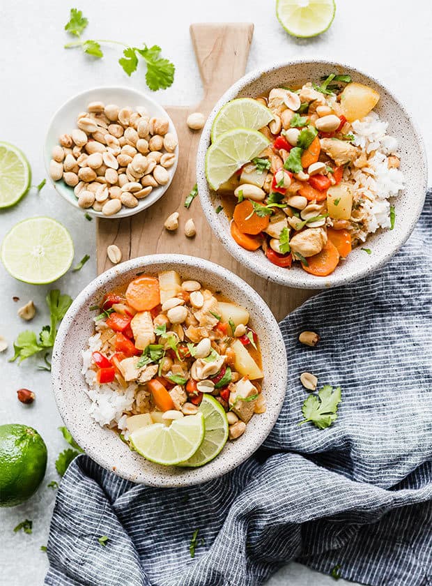 An overhead view of two bowls full of white rice and Massaman curry with lime wedges, cilantro, and peanuts for garnish.