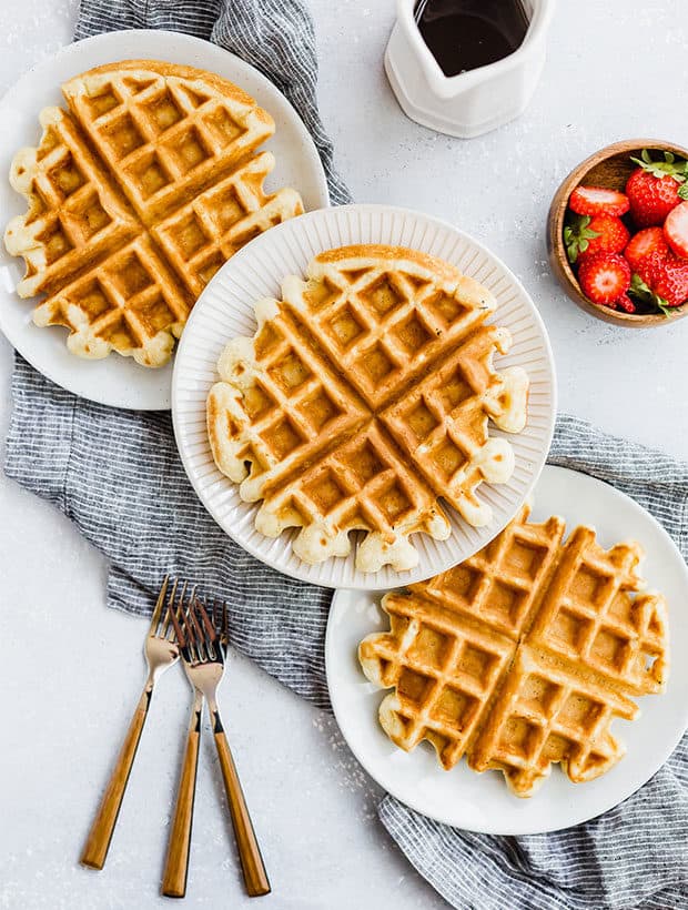 Three waffles on separate plates, with syrup in a white jar and a bowl of strawberries off to the side.