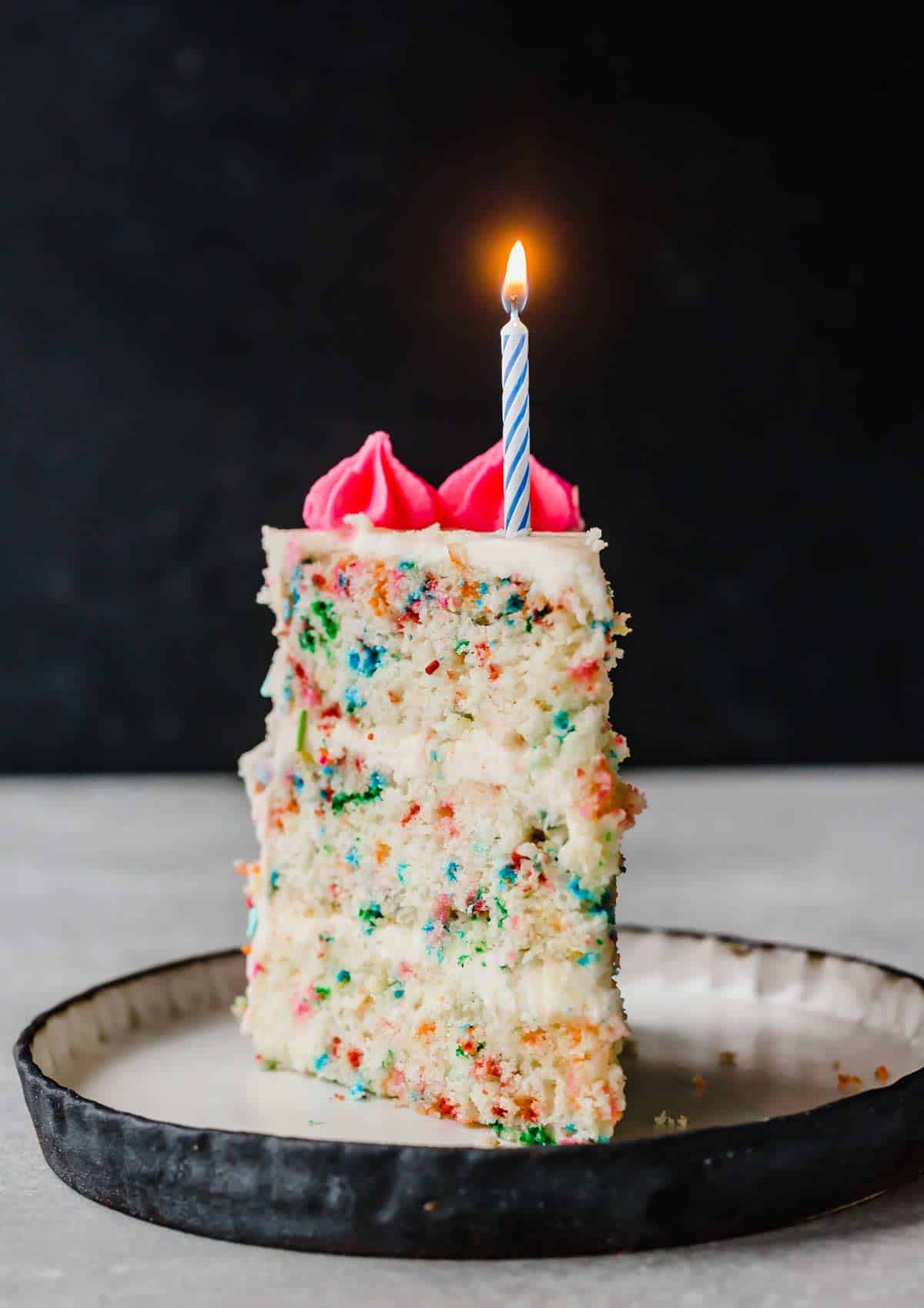 A slice of Funfetti Cake on a plate with a single blue candle lit on top of the slice.