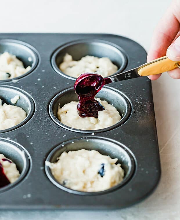 A muffin tin with blueberry muffin batter, and a fresh blueberry compote being poured into each muffin tin.