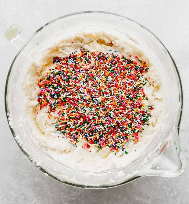 A glass mixing bowl full of Funfetti Cake batter with a pile of colorful sprinkles on the surface of the batter.