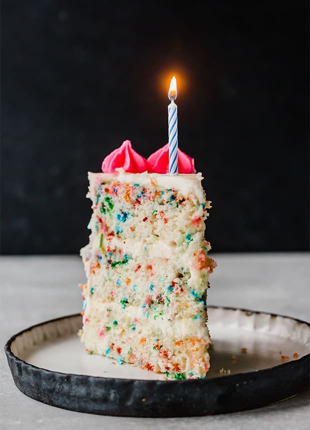 A slice of Funfetti Cake with one candle lit on the top of the cake slice.