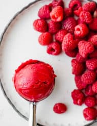 An ice cream scooper with a scoop of raspberry sorbet towards the left of the image, and a pile of red raspberries to the right of the scooper.