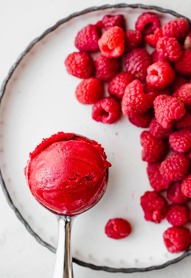 An ice cream scooper with a scoop of raspberry sorbet towards the left of the image, and a pile of red raspberries to the right of the scooper.