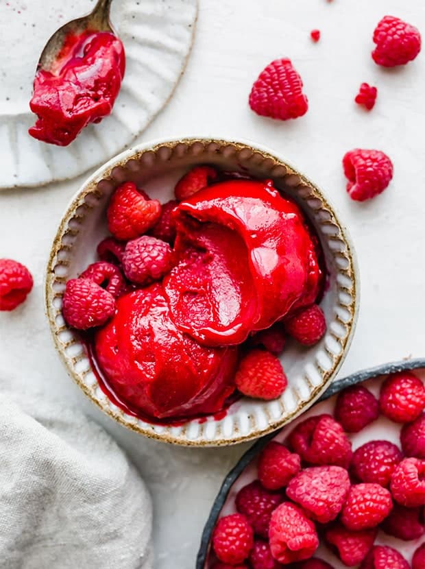 A bowl with two scoops of raspberry sorbet in the bowl; fresh raspberries scattered around the bowl.