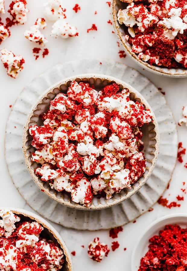 A bowl in the center of the photo full of red velvet popcorn with red velvet cake crumbs surrounding the bowl. 
