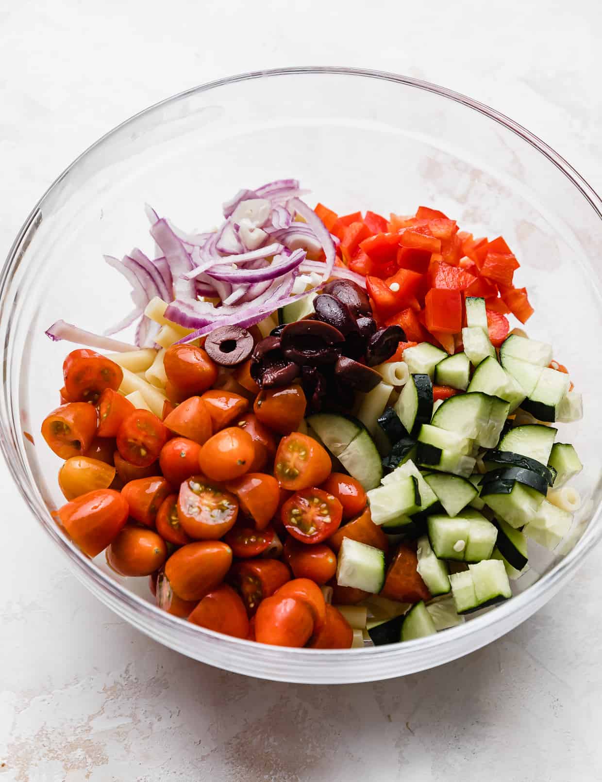 Penne noodles, tomatoes, kalamata olives, cucumbers, red onion, and red pepper in a glass bowl.