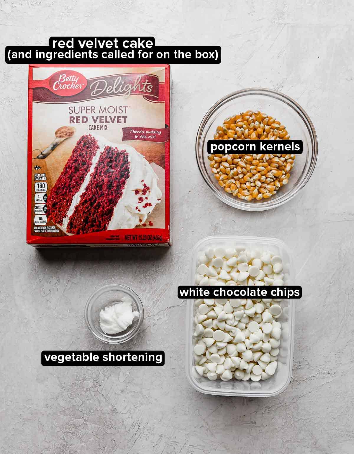 Red Velvet Popcorn ingredients on a light gray background: red velvet cake mix, popcorn kernels, white chocolate chips, and vegetable shortening.