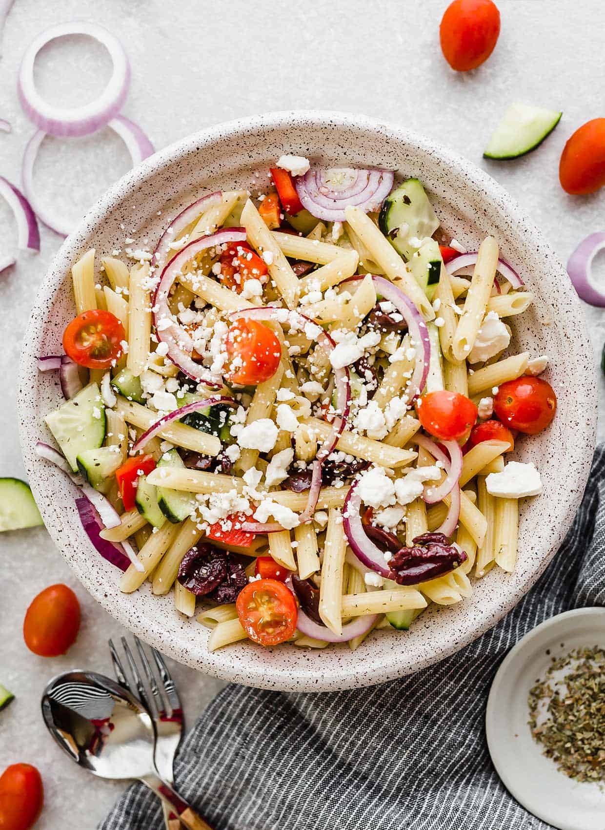 Greek pasta salad with penne pasta in a white bowl with tomatoes and red onion surrounding the bowl.