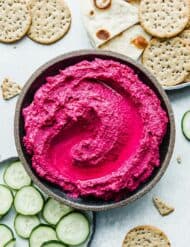Beet Hummus in a bowl surrounded by crackers, sliced cucumbers, and naan bread.