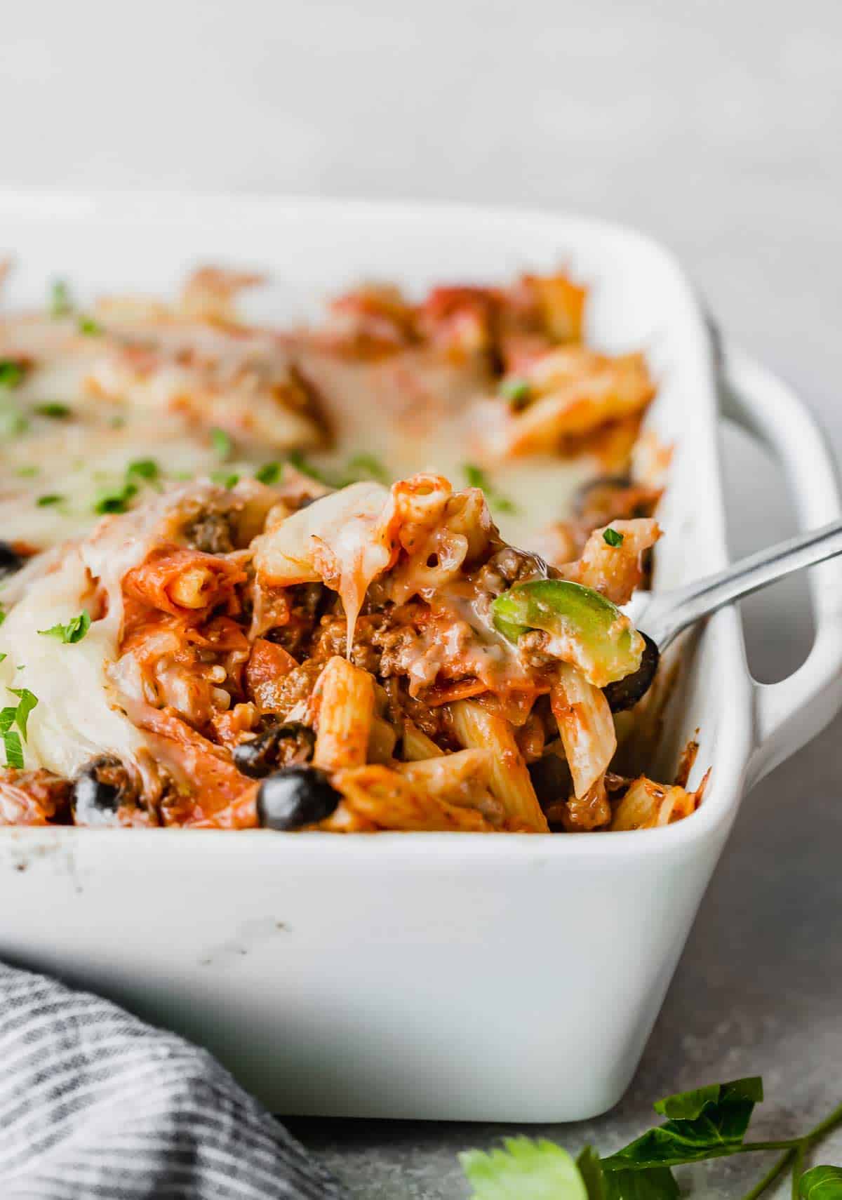 A spoon scooping up pizza casserole from a white baking dish.