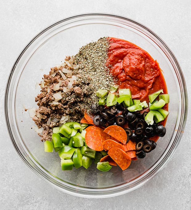 A glass bowl full of ingredients to make pizza casserole; ground meat, green bell peppers, pepperoni, marinara sauce, olives, and seasoning.