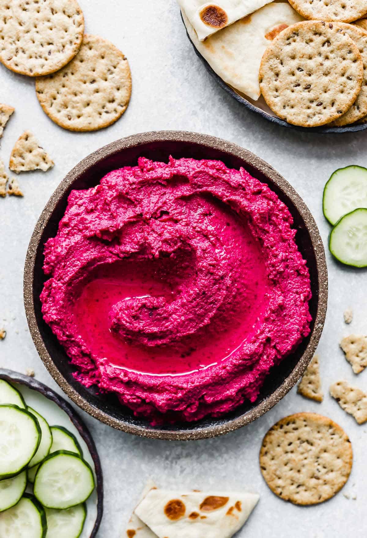 Beet Hummus on a black plate with sliced cucumbers and crackers surrounding the hummus.