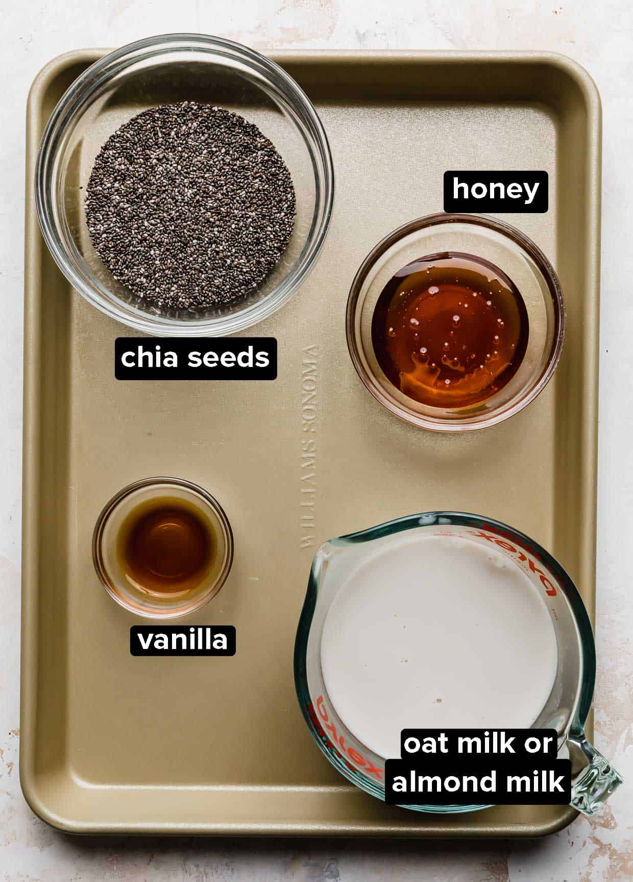 Chia Pudding with Oat Milk ingredients on a bronze baking sheet: chia seeds, vanilla, honey, and oat milk.