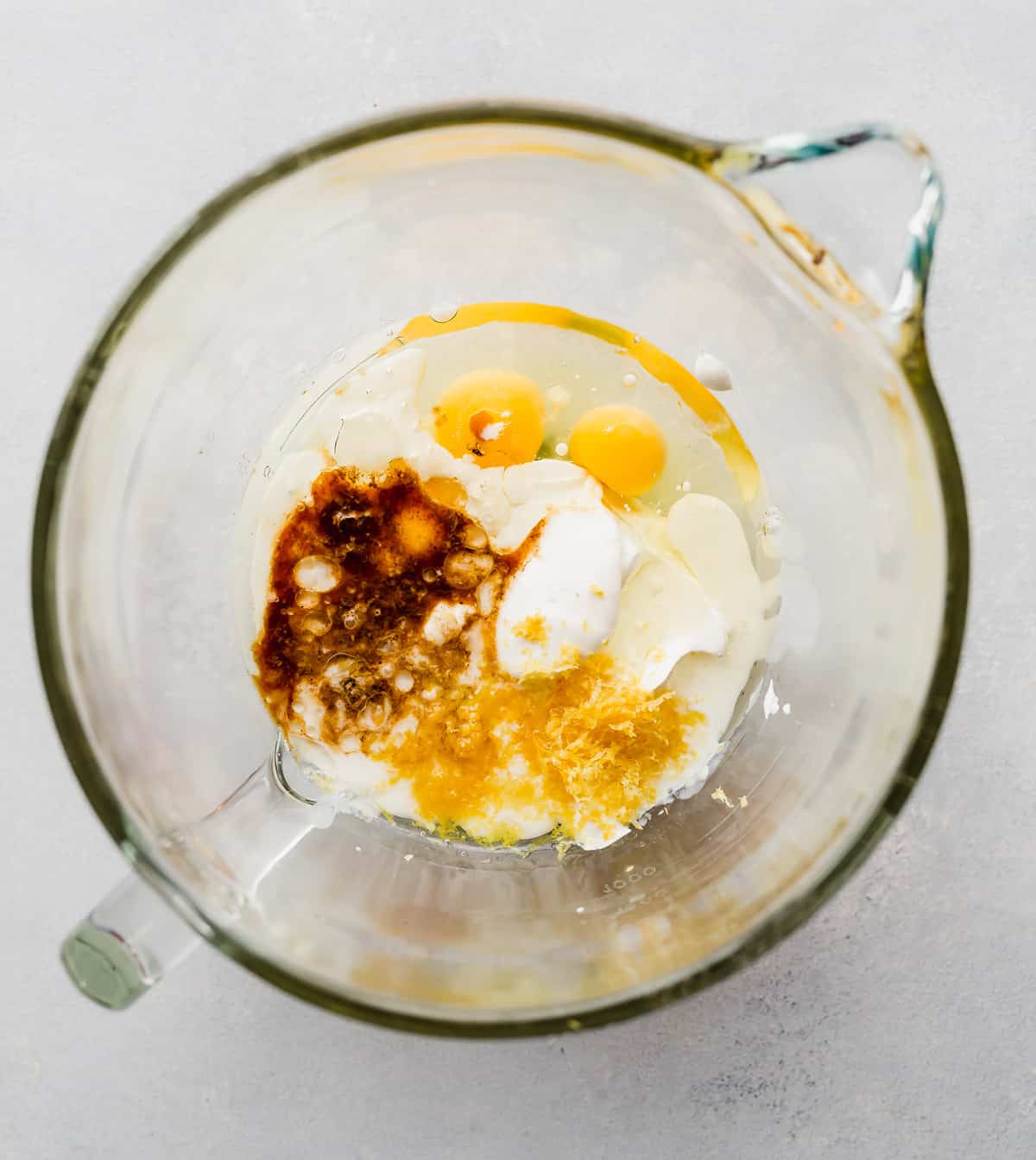 A glass mixing bowl with eggs, vanilla, sour cream, and lemon zest in it.