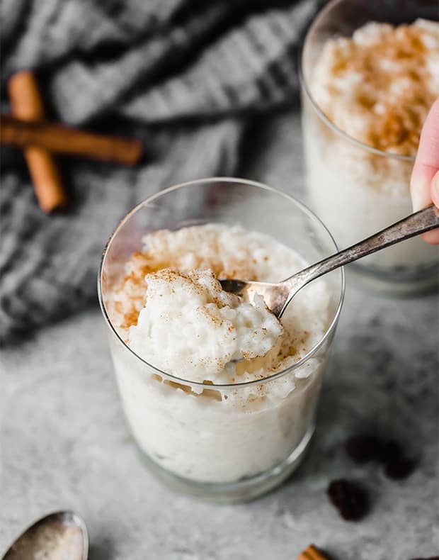 A glass cup full of Mexican Rice Pudding (Arroz con Leche) with a sprinkle of ground cinnamon over top, and a spoon scooping out a portion of the pudding.